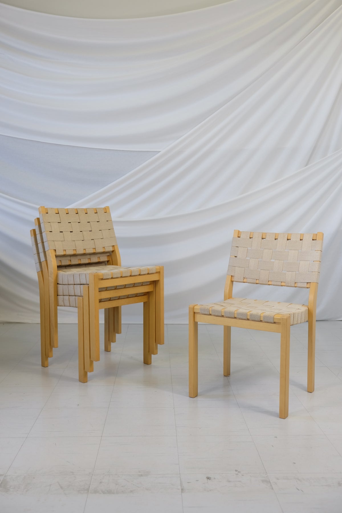 Set of four vintage Artek 611 chairs designed by Alvar Aalto with birch frames and linen webbing, visible wear and staining, photographed in front of a white fabric backdrop.